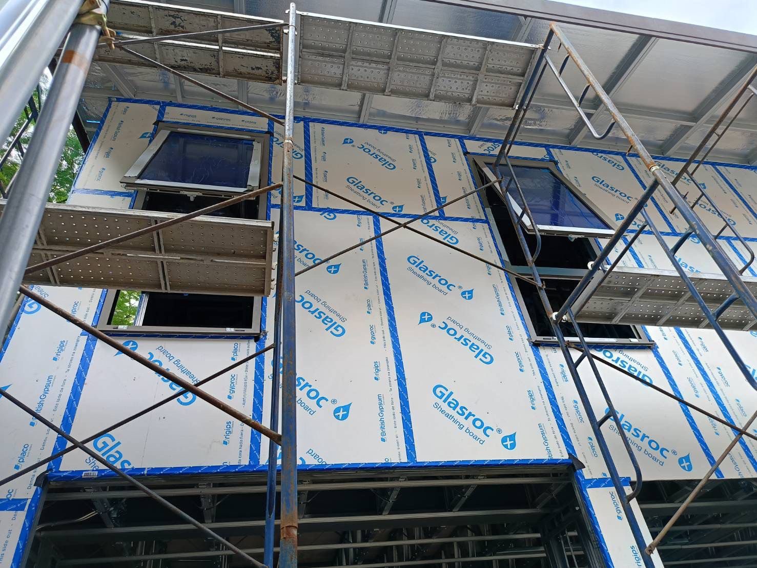 A multi-storey wall under construction: white Glasroc X boards with blue tape on every joint and the windows fitted, behind scaffolding