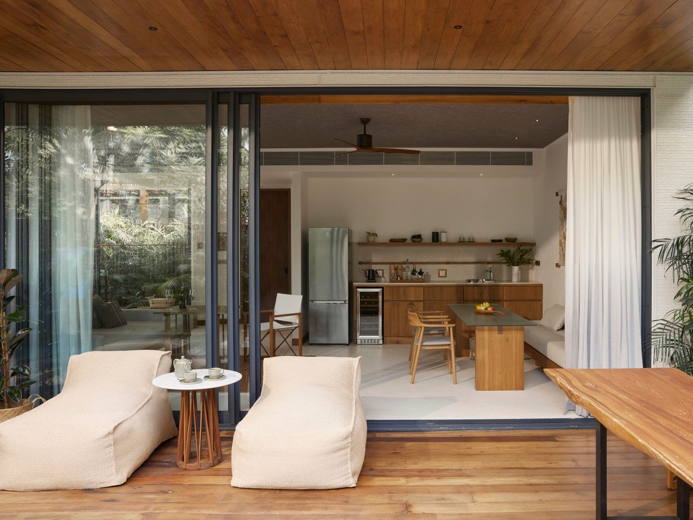 A timber terrace at a pool villa, its dark-framed sliding glass doors opened wide onto the kitchen and dining table