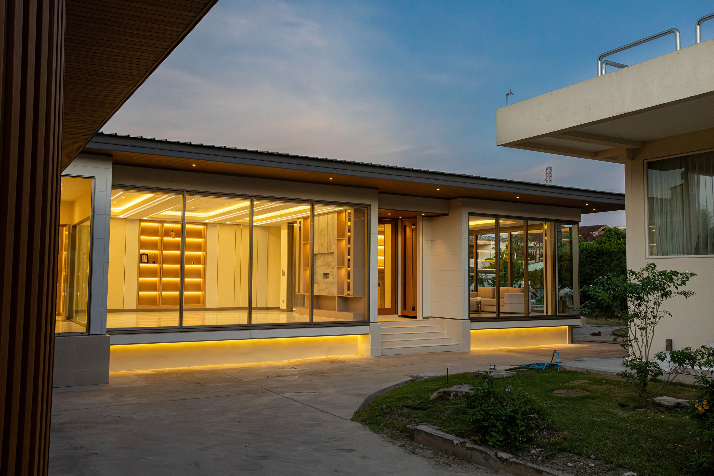 A single-storey house at dusk, its large glass wall lit from inside, seen across the front lawn