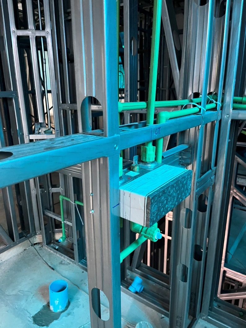 Green water pipes run through the oval holes punched in the steel studs to a fitting fixed in the frame, with blue drain pipes rising from the floor