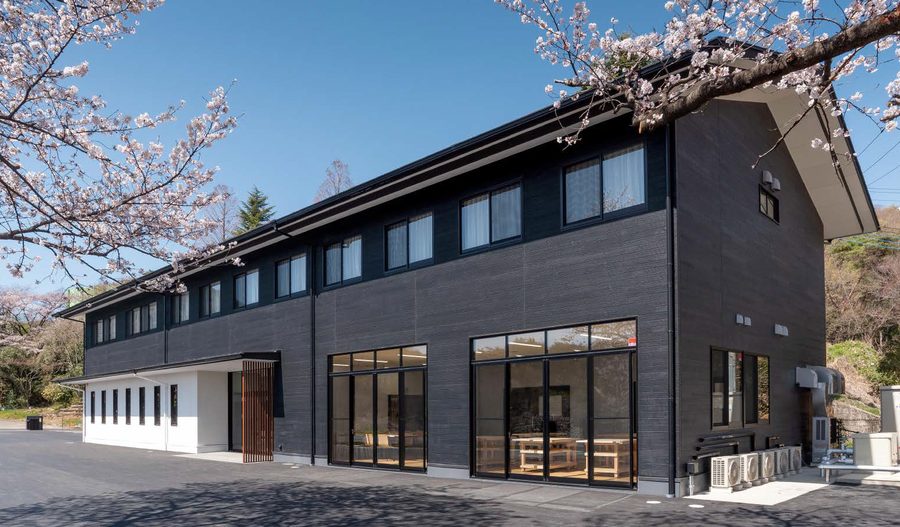 A two-storey school building in Japan clad in black wood-look panels, with tall glazed doors, a white entrance wing and cherry blossom overhead