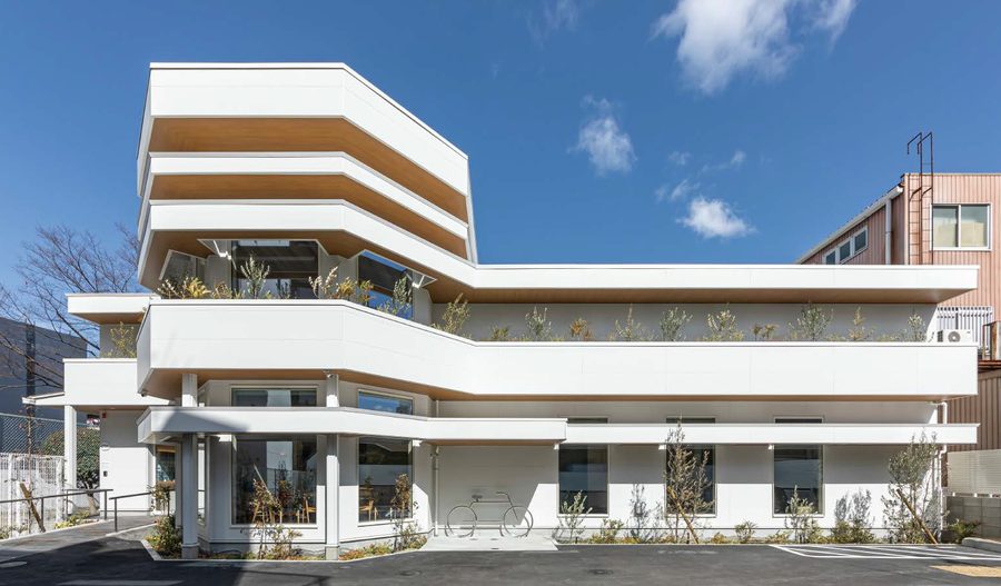 A white clinic in Japan built up in stacked horizontal bands, with wood-toned soffits and planted terraces