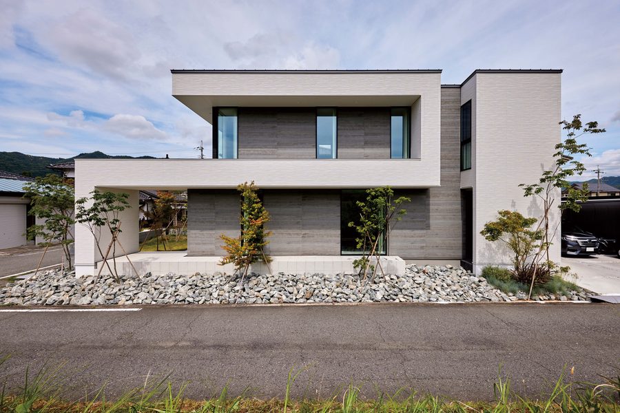 A flat-roofed modern house in Japan: a white cantilevered frame over grey wood-look walls, behind a bed of rocks and young maples