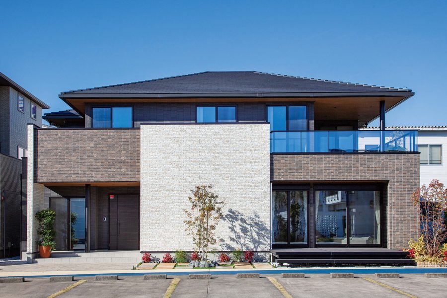 A two-storey house in Japan with a white stacked-stone-look feature wall between brown brick-look walls, a glass balcony and deep eaves