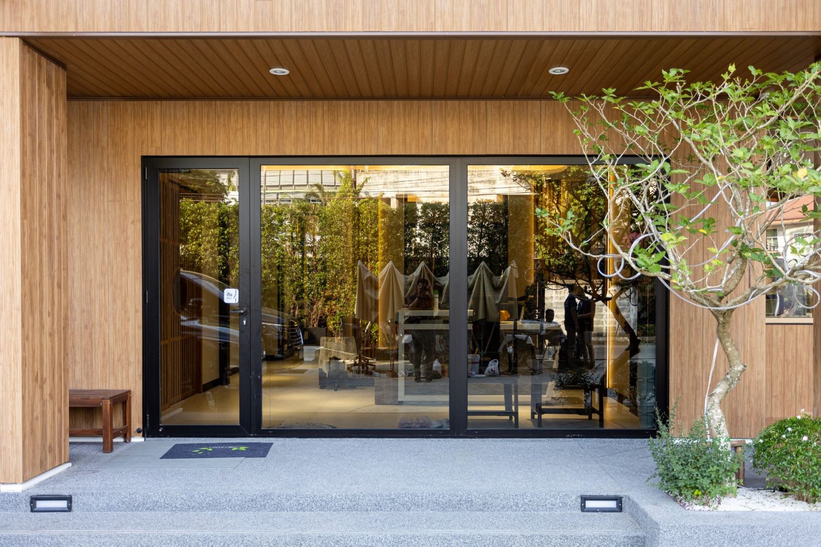 An office entrance: light wood-grain walls and soffit, black-framed glass doors, a tree and path lights in front