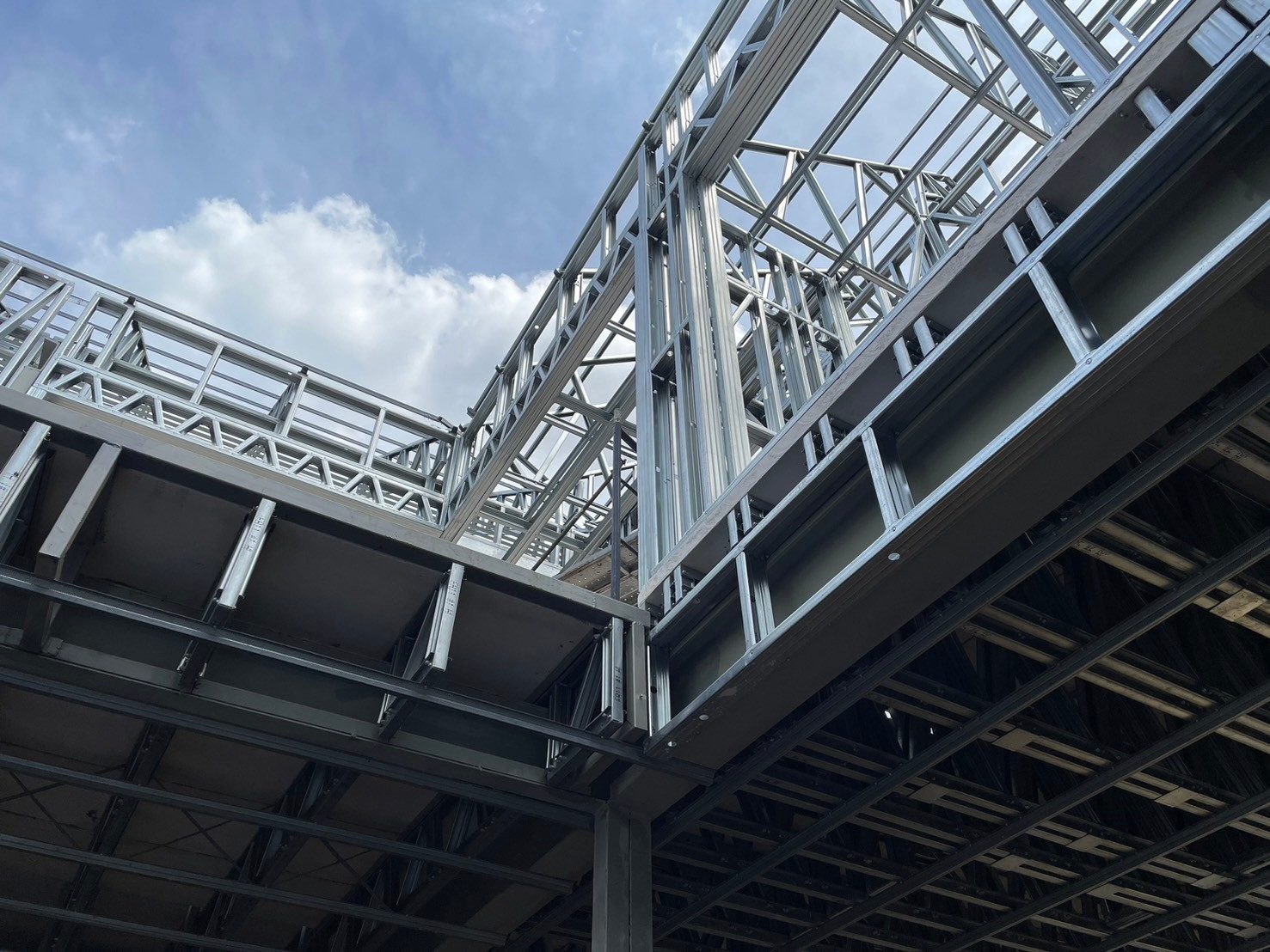 Looking up past steel floor joists to the upper walls and roof frame of a light gauge steel house against a blue sky