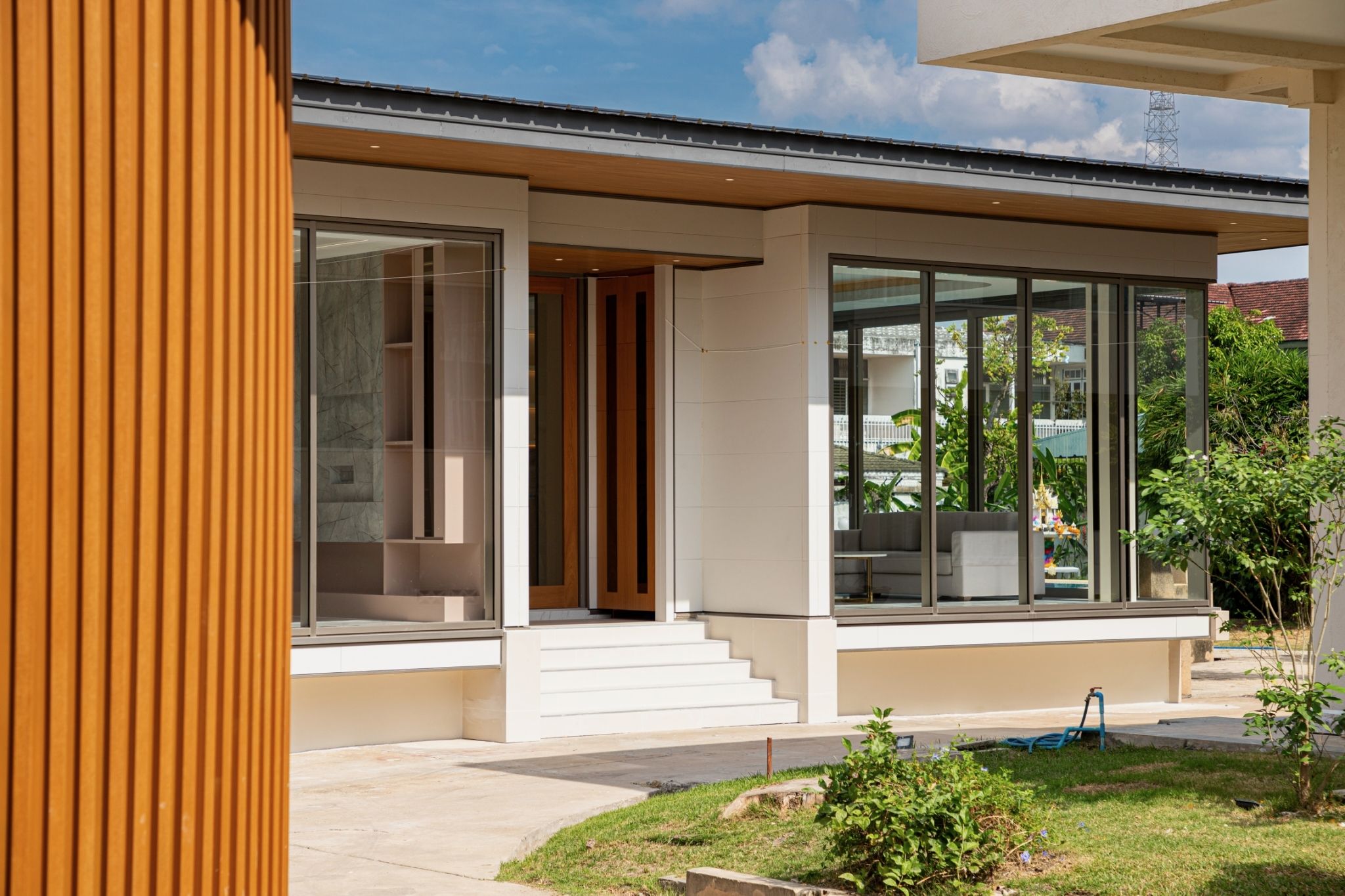 The entrance side of a single-storey house in daylight: glass walls in slim frames, a timber-look door and steps under a deep eave