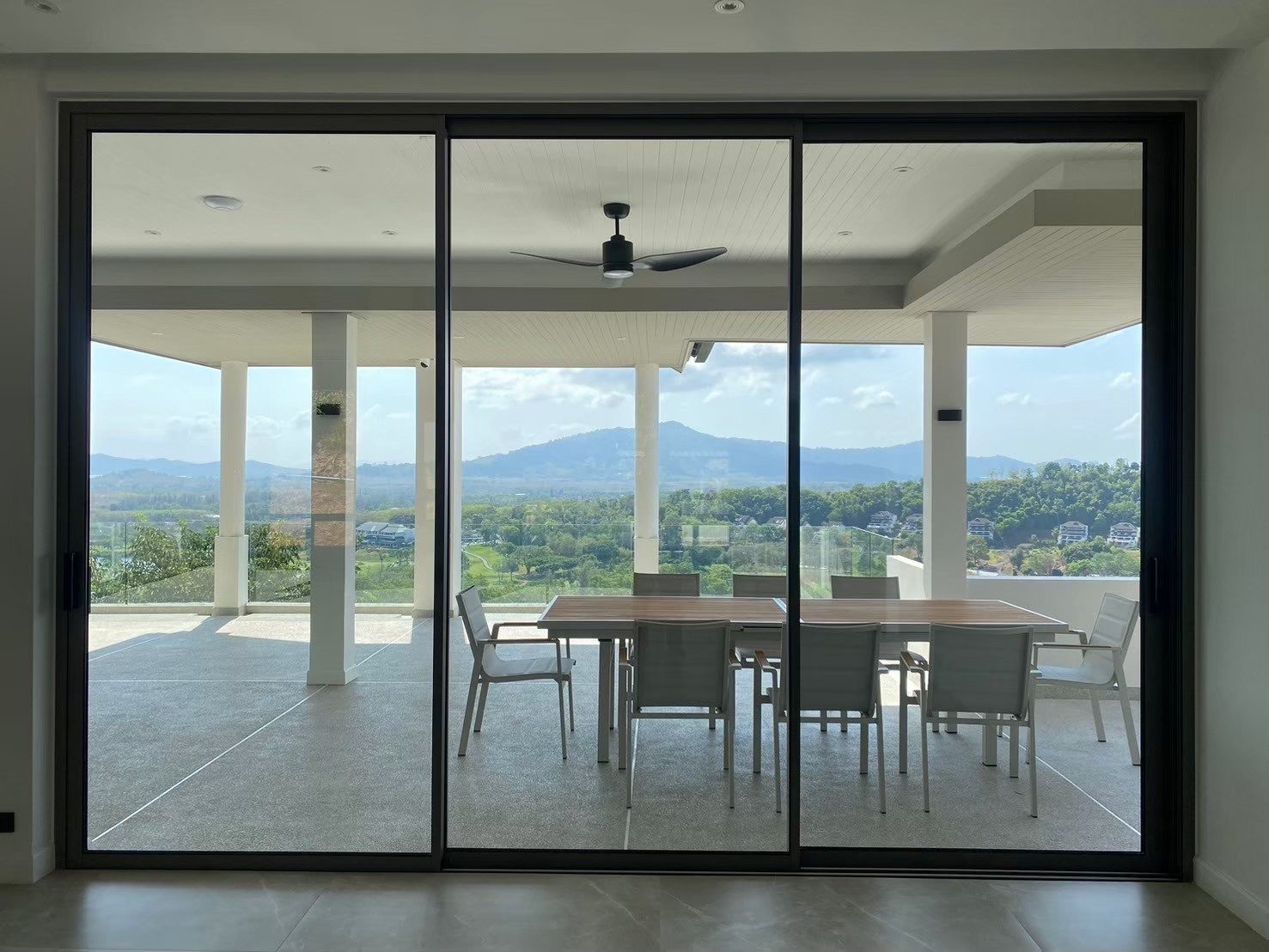 A three-panel Technal SOLEAL GYn sliding door in dark frames looks out to a covered terrace with a dining table and green hills beyond