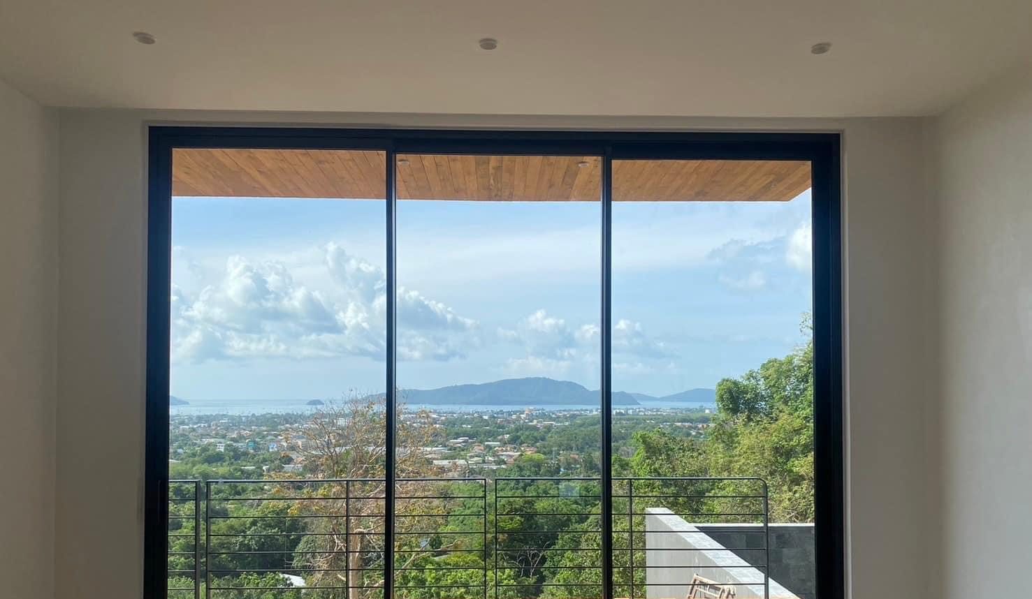 A three-panel Technal SOLEAL GYn sliding door in black frames opens a white room onto a timber deck and a view over the sea and islands