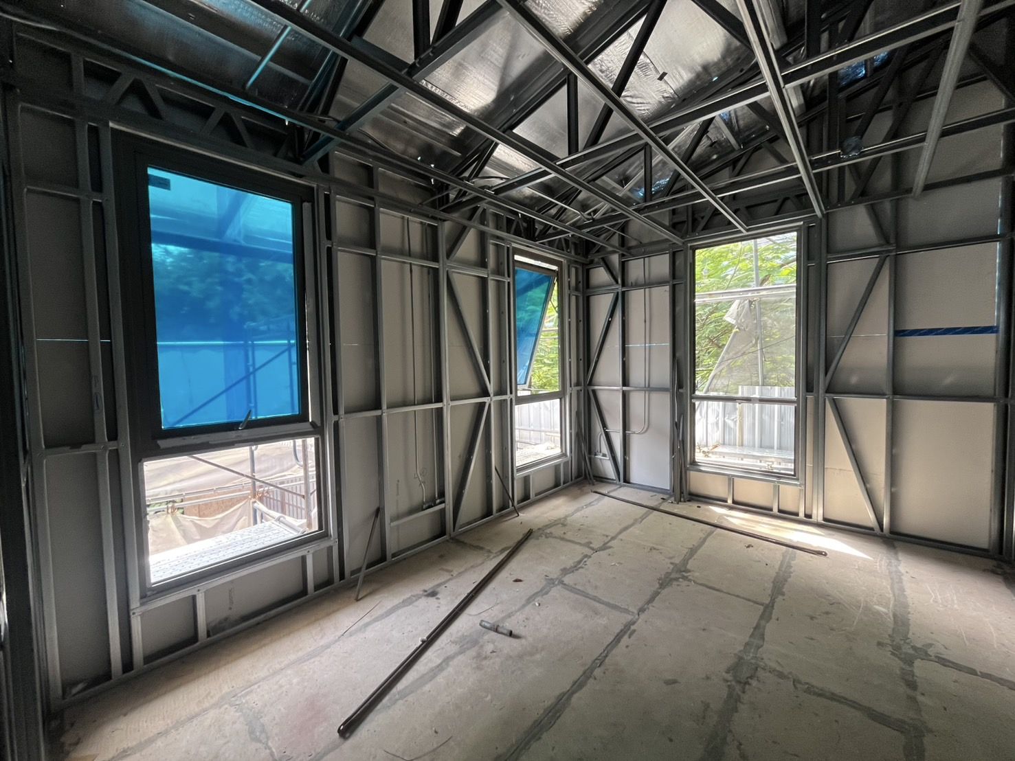 Inside a steel-framed house: aluminium windows, still in their blue film, fitted into openings in the light gauge steel frame, with bracing and roof trusses around them